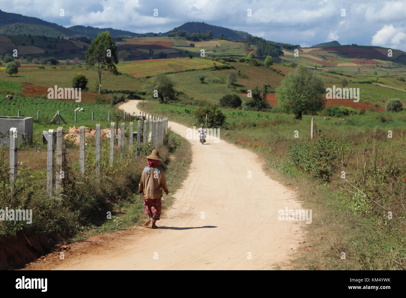 Eine Landschaft Bild eines lokalen Frau in traditioneller Kleidung weg von der Kamera zu Fuß auf einem staubigen Schotterstraße in Myanmar, die Straße schlängelt sich zwischen Hügeln und weit Stockfoto