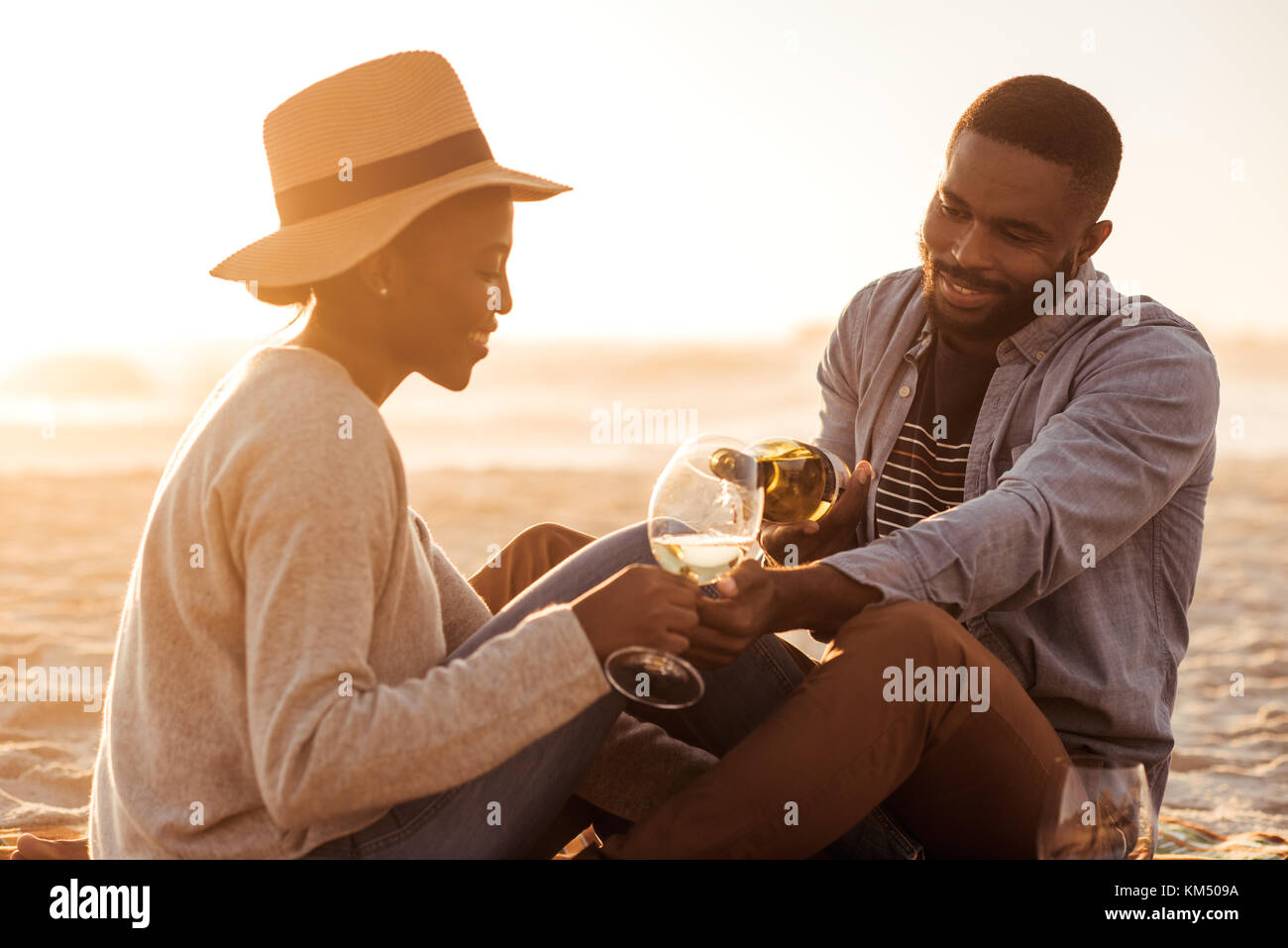 Lächelnd afrikanischen Paar Wein trinken und am Strand bei Sonnenuntergang Stockfoto