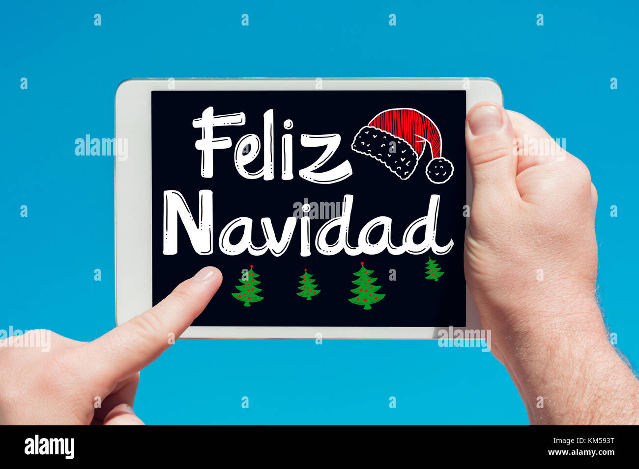 Mann hält ein Tablet-gerät mit Text in Spanisch "Feliz Navidad (Frohe Weihnachten) und berühren Sie den Bildschirm mit einem Finger mit blauen Himmel im Hintergrund. Stockfoto