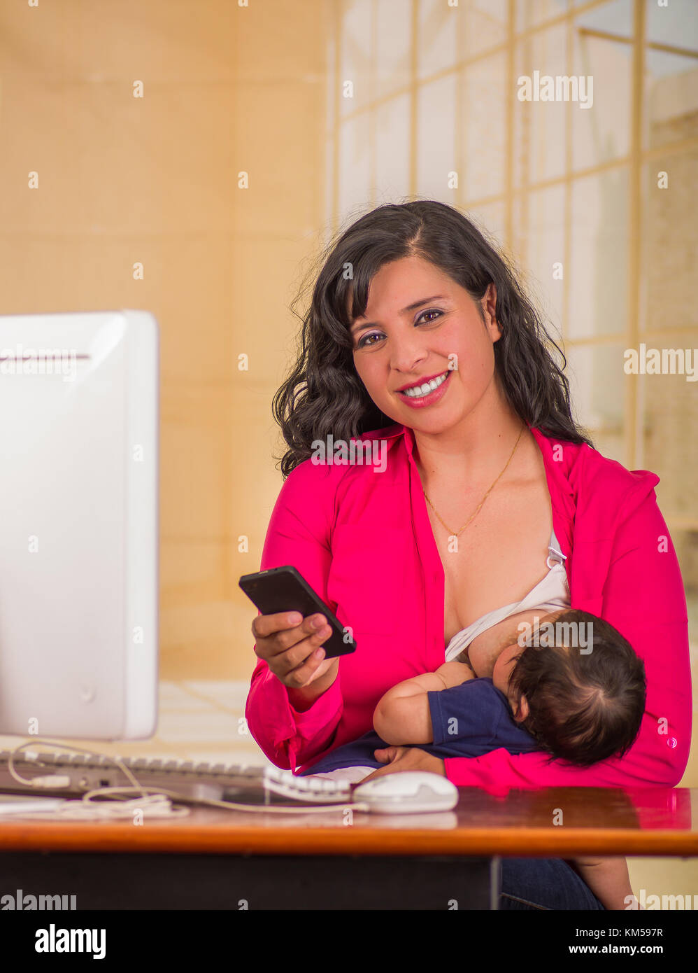 Junge schöne Geschäftsfrau, die im Büro sitzend mit seinem baby boy an ihrem Arbeitsplatz, stillen, während sie mit ihrem Handy in einem unscharfen Hintergrund ist Stockfoto