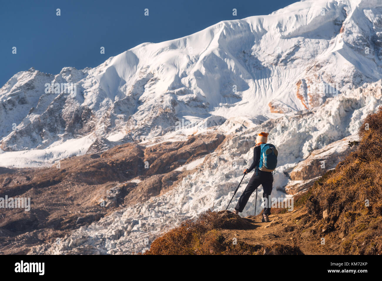 Stehende junge Frau mit Rucksack auf dem Berg und Blick auf die Berge und den Gletscher bei Sonnenuntergang. Landschaft mit Mädchen, Felsen mit Schnee Stockfoto
