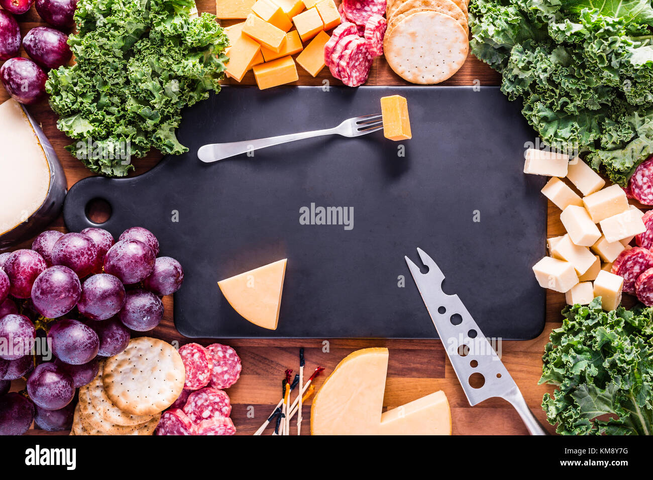 Fleisch- und Käseplatte mit einem schwarzen Brett. Verschiedene Käse und Wurst werden mit Grünkohl und lila Trauben angezeigt. Stockfoto
