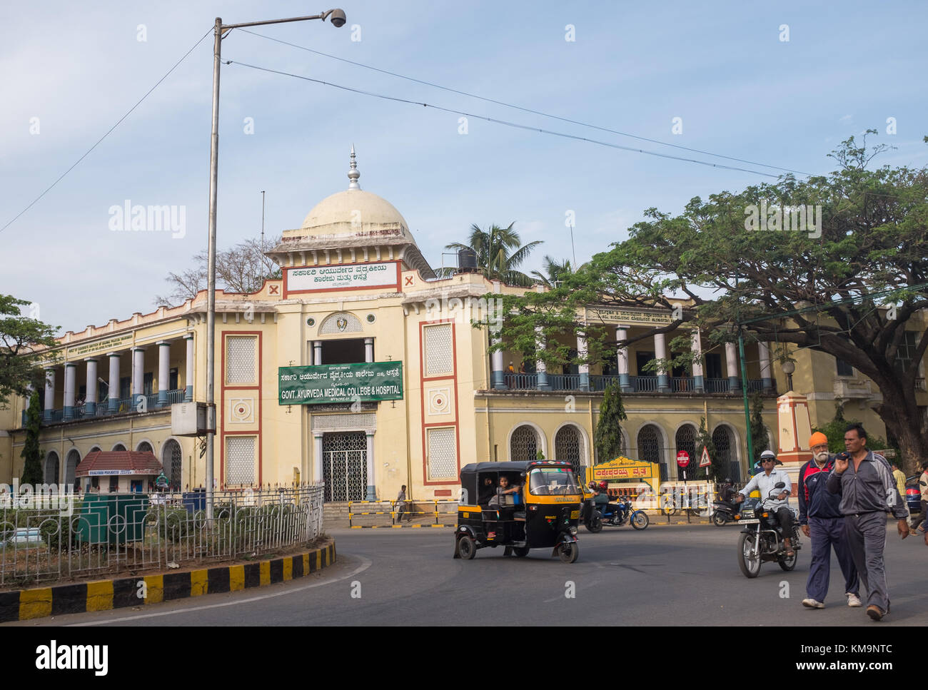 Die Außenseite des Regierung ayurveda Medical College und Krankenhaus, Menschen zu Fuß und das Fahren von Fahrzeugen auf der Straße im Vordergrund Mysore, mysuru, Karnataka, Stockfoto