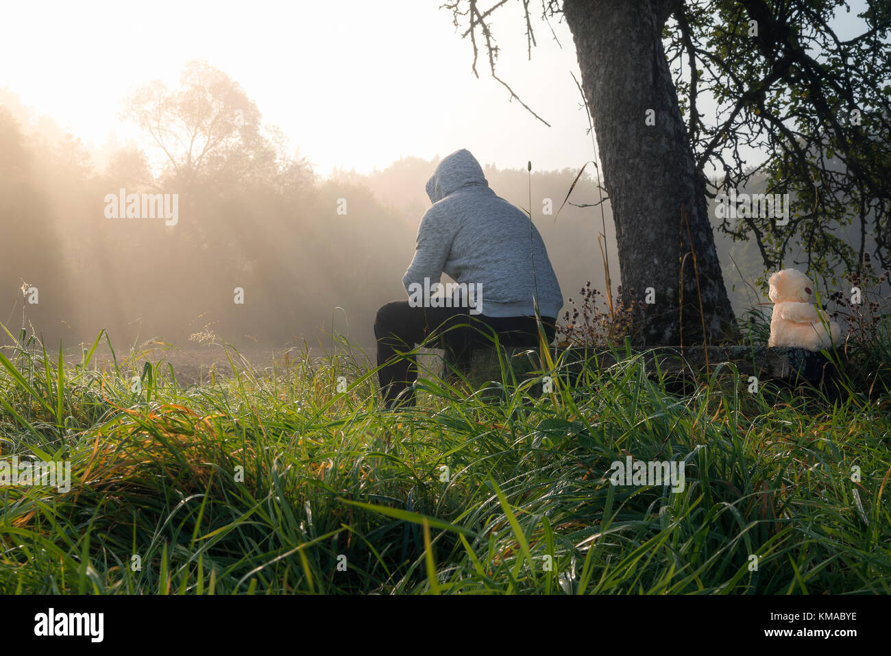 Beziehung Themen Bild mit einem Mann und einem Teddybär sitzt auf einer Holzbank, unter einem Baum, beim Genießen der Morgennebel und Sonnenstrahlen. Stockfoto