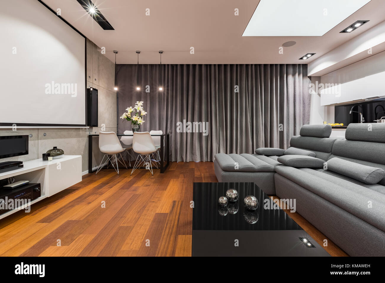Grau-weißes Apartment mit Projektionsleinwand, Sofa, Esstisch und Kochnische Stockfoto