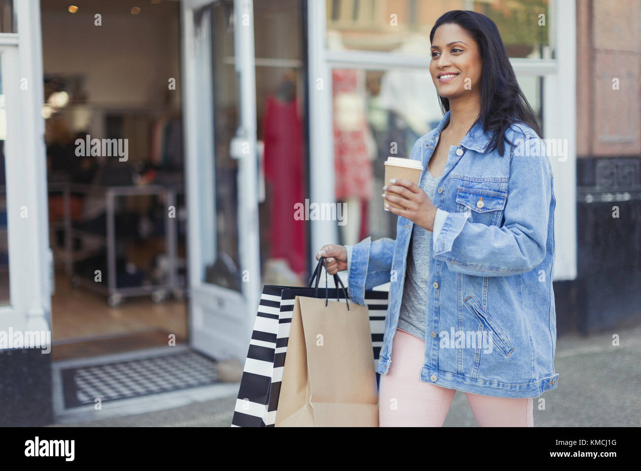 Lächelnde Frau, die mit Kaffee und Einkaufstaschen am Schaufenster entlang geht Stockfoto