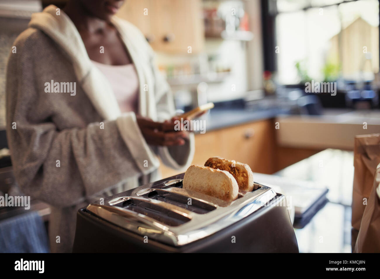Frau schreibt mit Smartphone, Toasten von Brot im Toaster in der Küche Stockfoto