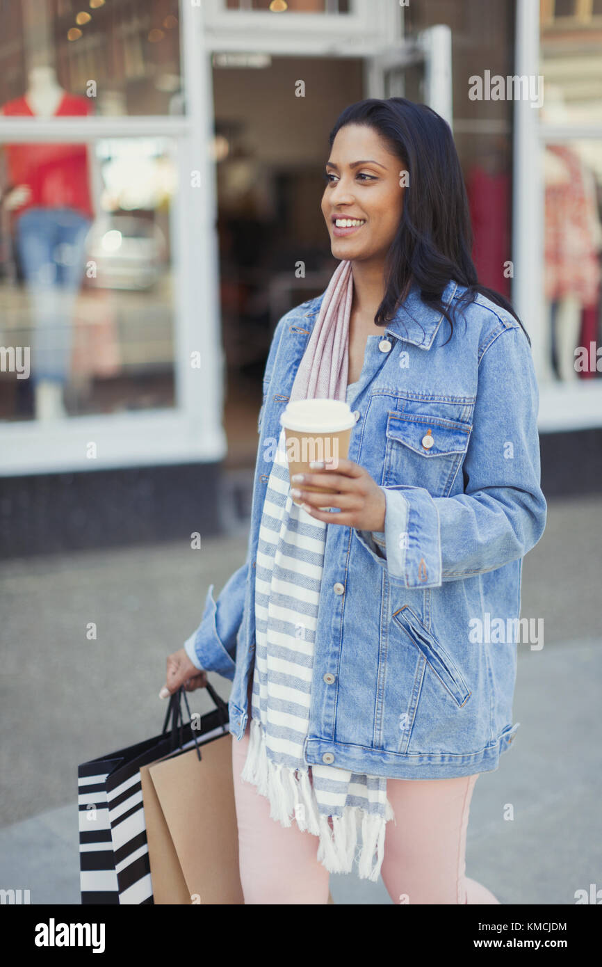 Lächelnde Frau, die mit Kaffee und Einkaufstaschen am Schaufenster entlang geht Stockfoto