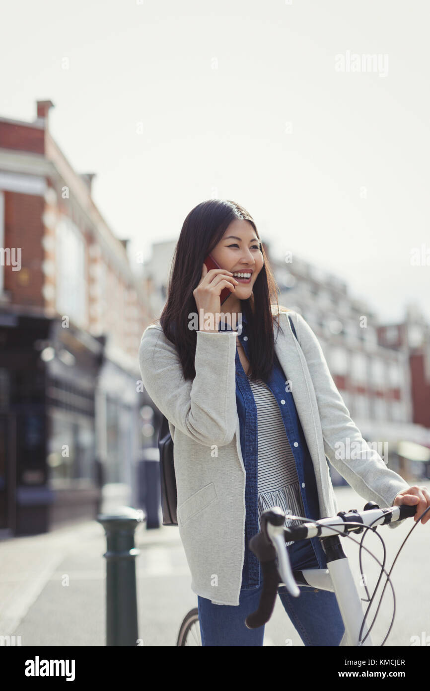 Lächelnde junge Frau pendelt mit dem Fahrrad, spricht auf dem Handy auf der sonnigen städtischen Straße Stockfoto