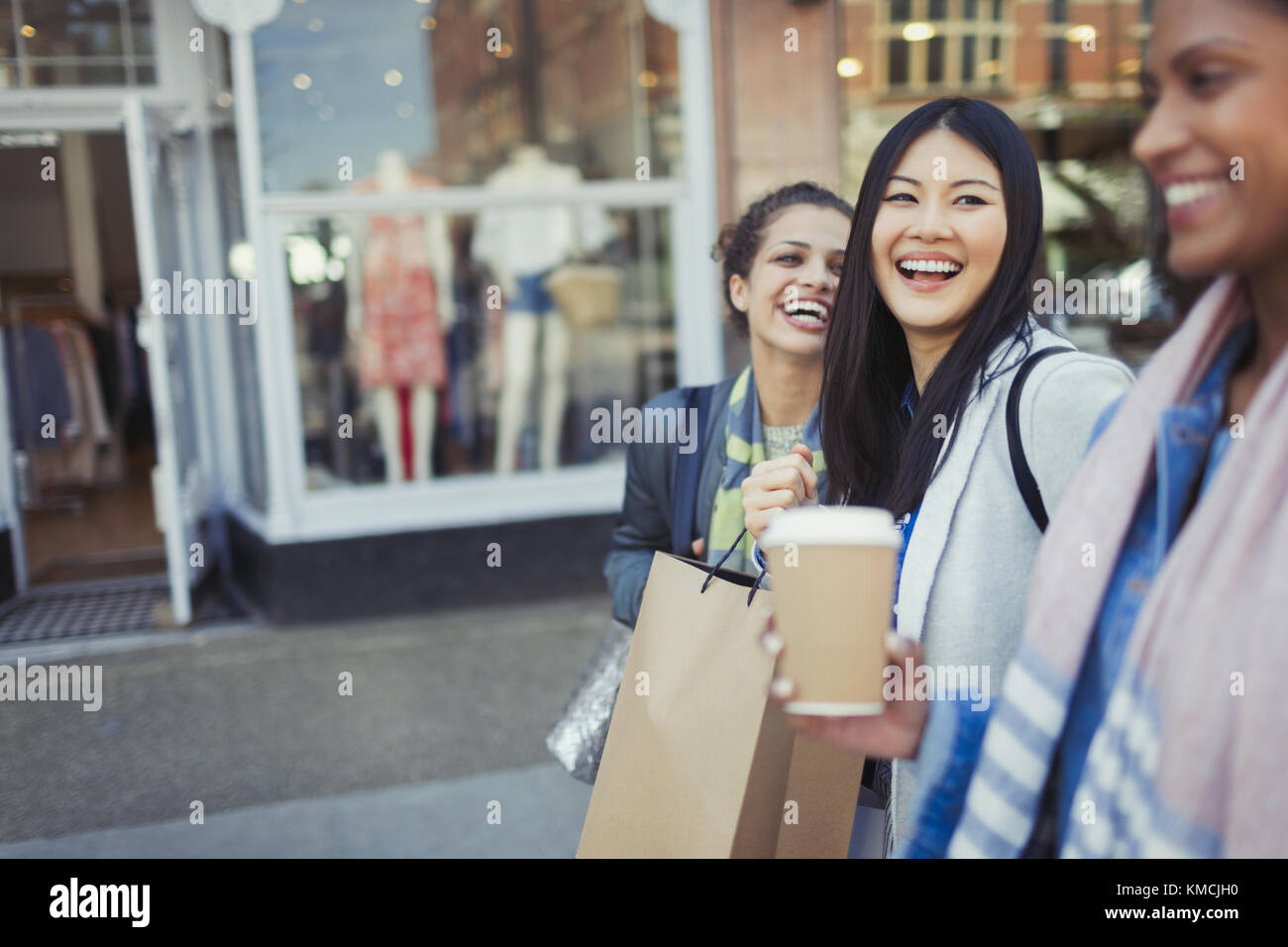 Lachende Freundinnen, die mit Kaffee und Einkaufen entlang der Ladenfront gehen Taschen Stockfoto