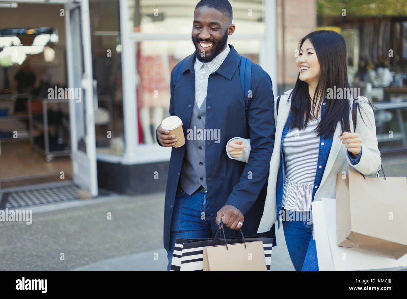 Lächelndes junges Paar, das Arm in Arm entlang der Schaufenster mit geht Kaffee und Einkaufstaschen Stockfoto