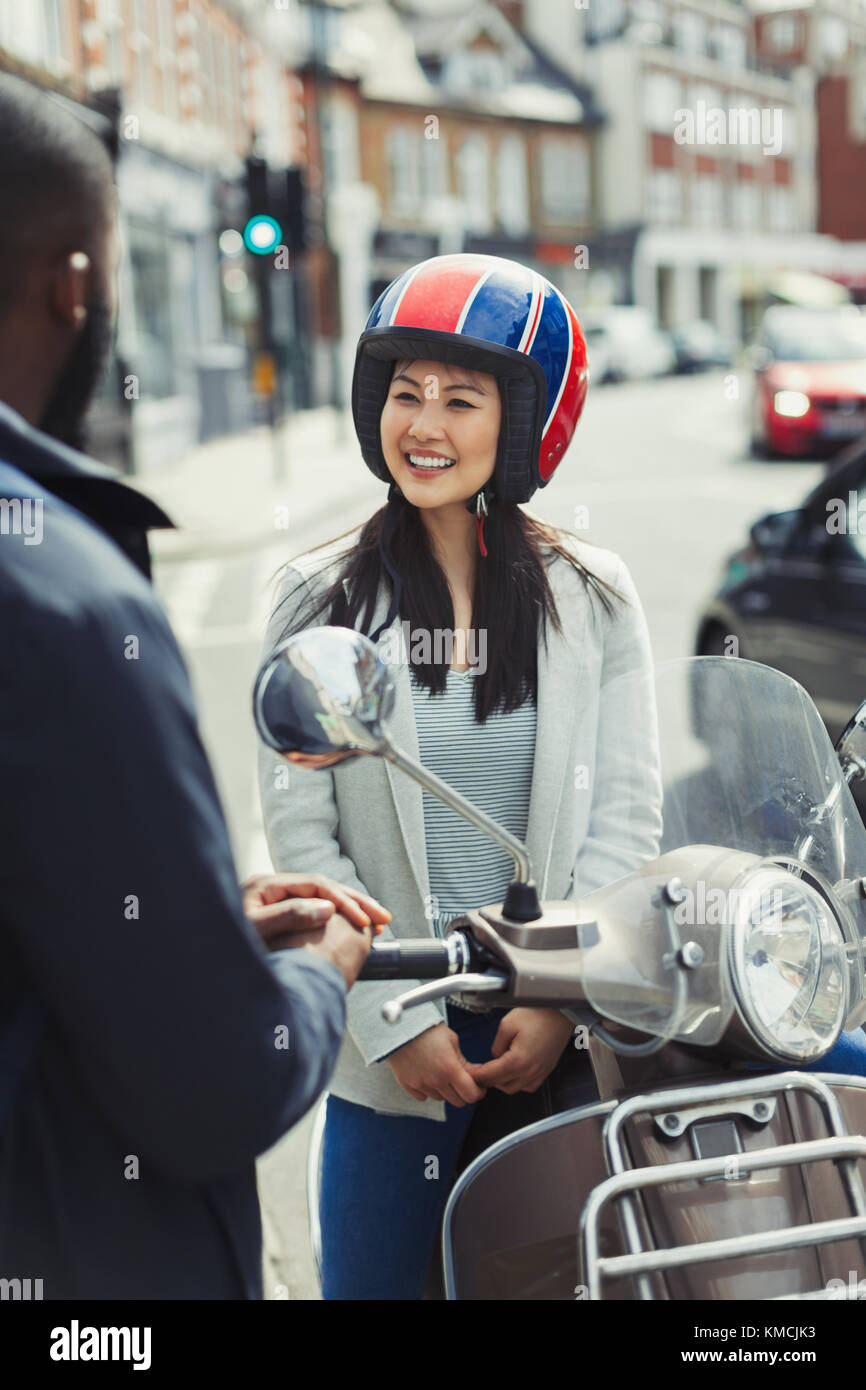 Lächelnde junge Frau im Helm auf Motorroller, im Gespräch mit einem Freund auf der städtischen Straße Stockfoto