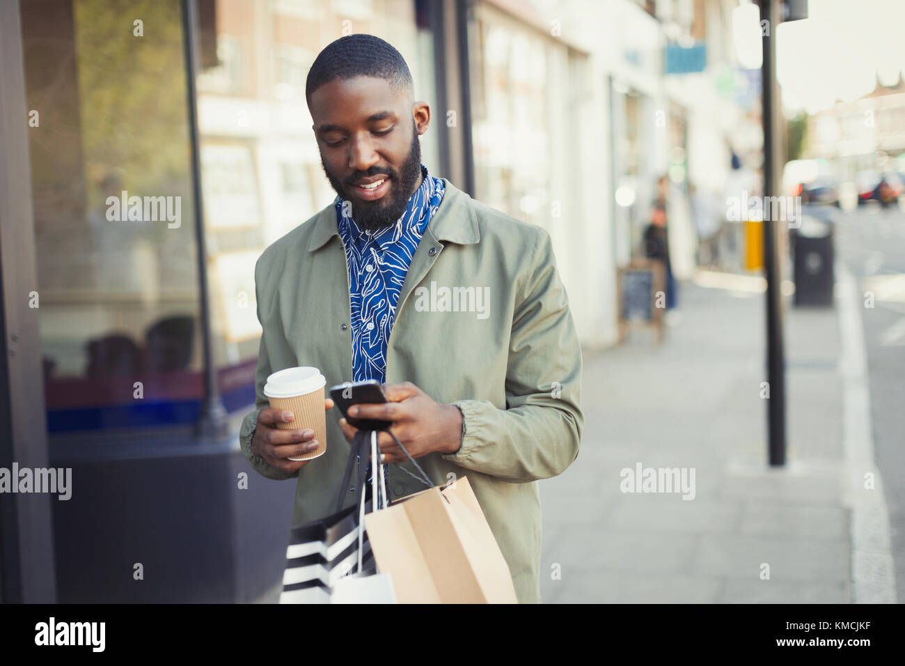 Junger Mann mit Kaffee und Einkaufstaschen SMS mit Zelle Telefon auf städtischen Bürgersteig Stockfoto