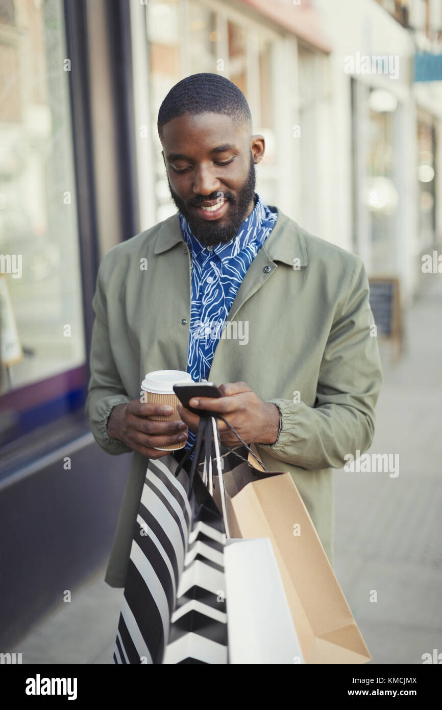 Lächelnder junger Mann mit Kaffee und Einkaufstaschen mit SMS Handy auf dem städtischen Bürgersteig Stockfoto
