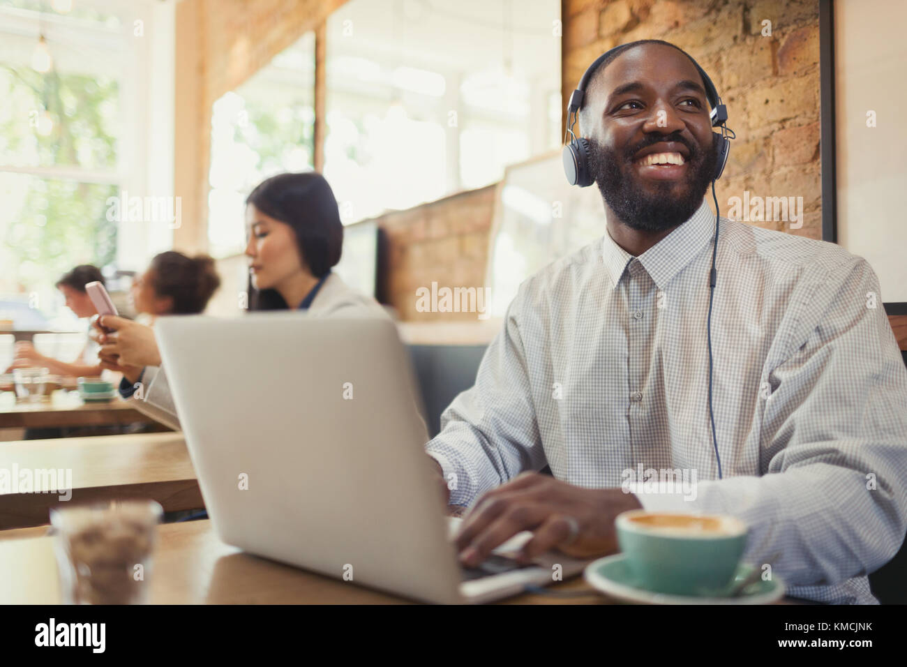 Lächelnder junger Mann mit Kopfhörern, der Laptop benutzt und Kaffee trinkt Am Kaffeetisch Stockfoto