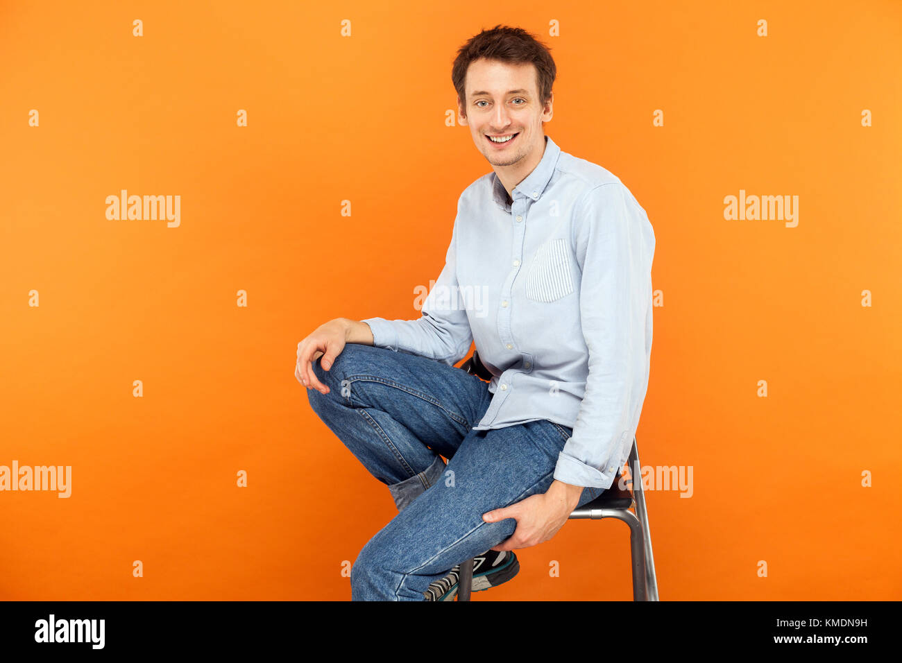 Glück Mann sitzen auf einem Stuhl, an der Kamera und toothy Lächeln suchen. Studio shot, orange Hintergrund Stockfoto