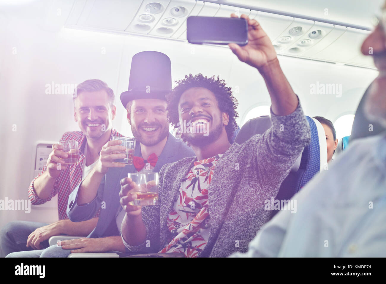 Enthusiastische junge männliche Freunde mit Kamera-Handy trinken und nehmen Selfie im Flugzeug Stockfoto