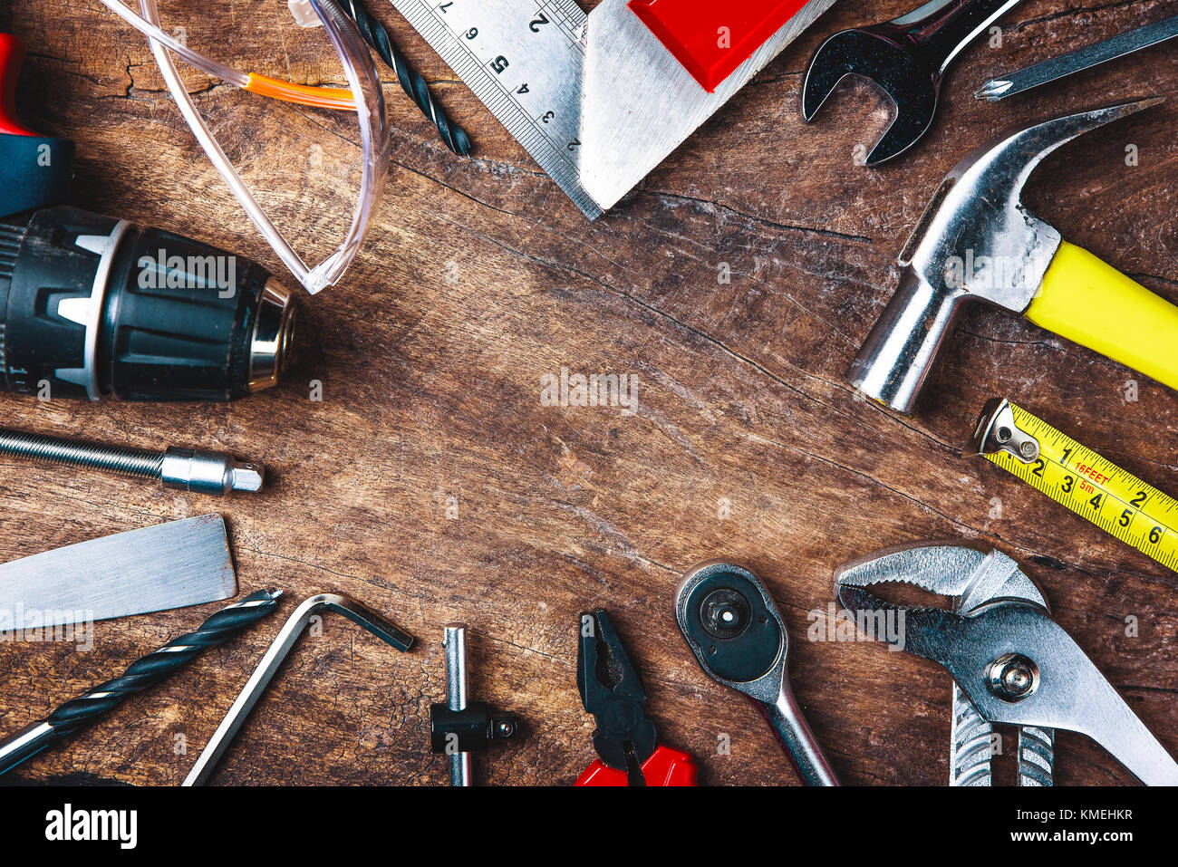 Blick von oben auf die Werkzeuge, Schraubendreher, Schraubenschlüssel, Hammer, Schraubenzieher, Zange, elektrische Bohrmaschine, Maßband, maschinist Square und Schutzbrille auf Holz zurück Stockfoto