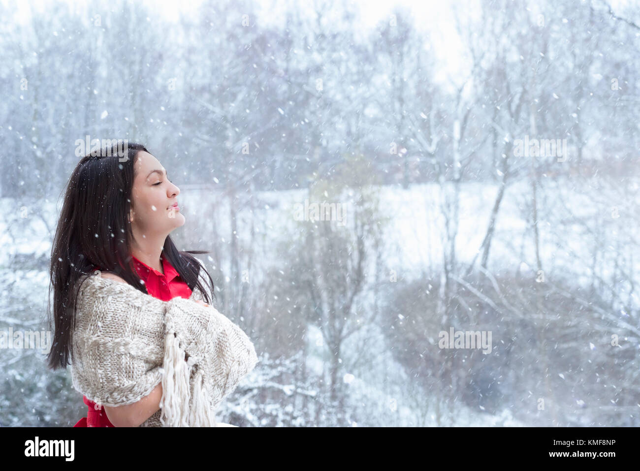 Attraktive junge Frau mit langen dunklen Haaren, im roten Kleid, mit einem handgefertigten Schal bedeckt, mit geschlossenen Augen, geniessen die Schneefall. Stockfoto