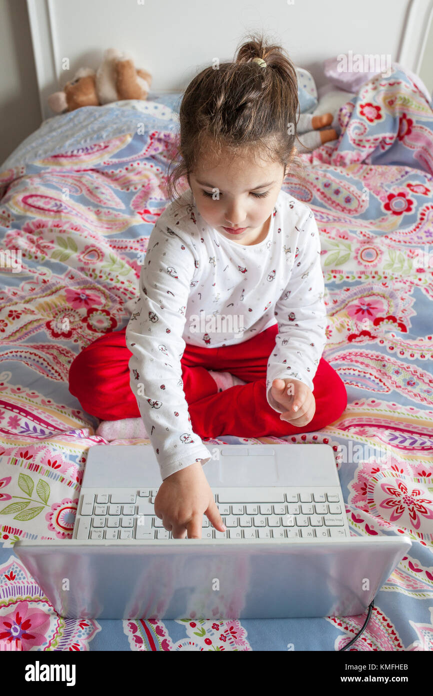 Kleines Mädchen im Bett sitzen und spielen online Spiele mit Laptop. Sie ist durch Berühren des Bildschirms Stockfoto