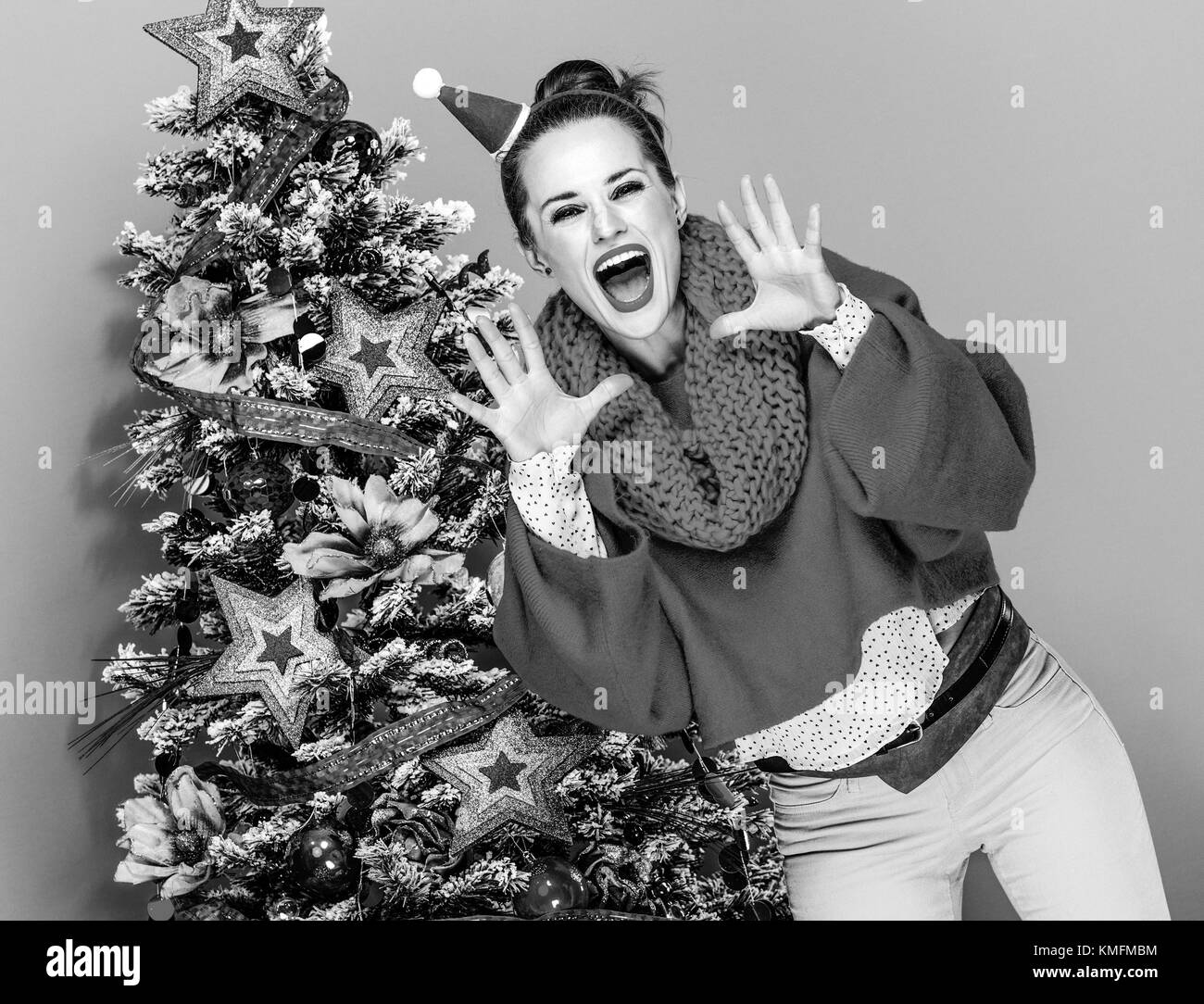 Festliche Jahreszeit. lächelnd trendy Frau in bunten Kleidern in der Nähe von Weihnachten Baum auf gelben Hintergrund Schreien über Megaphon geformten Hände Stockfoto