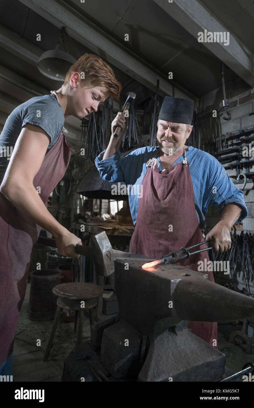 Schmied und Lehrling hammerten in der Werkstatt eine rote Bar mit heißem Eisen Stockfoto