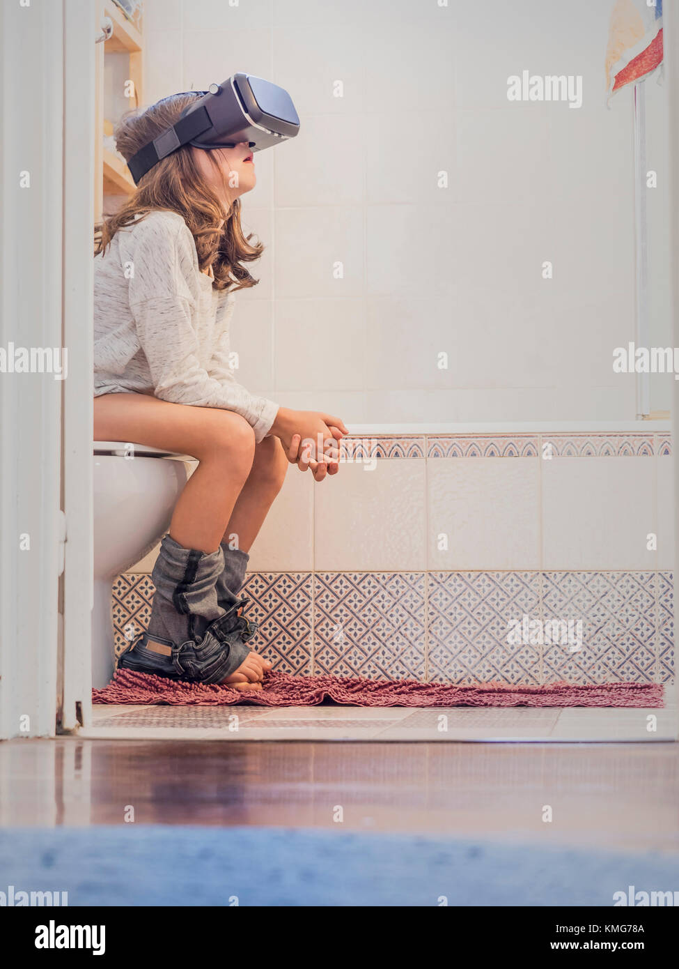 Mädchen, das auf einer Toilettenschüssel sitzt und eine Virtual-Reality-Brille trägt Stockfoto