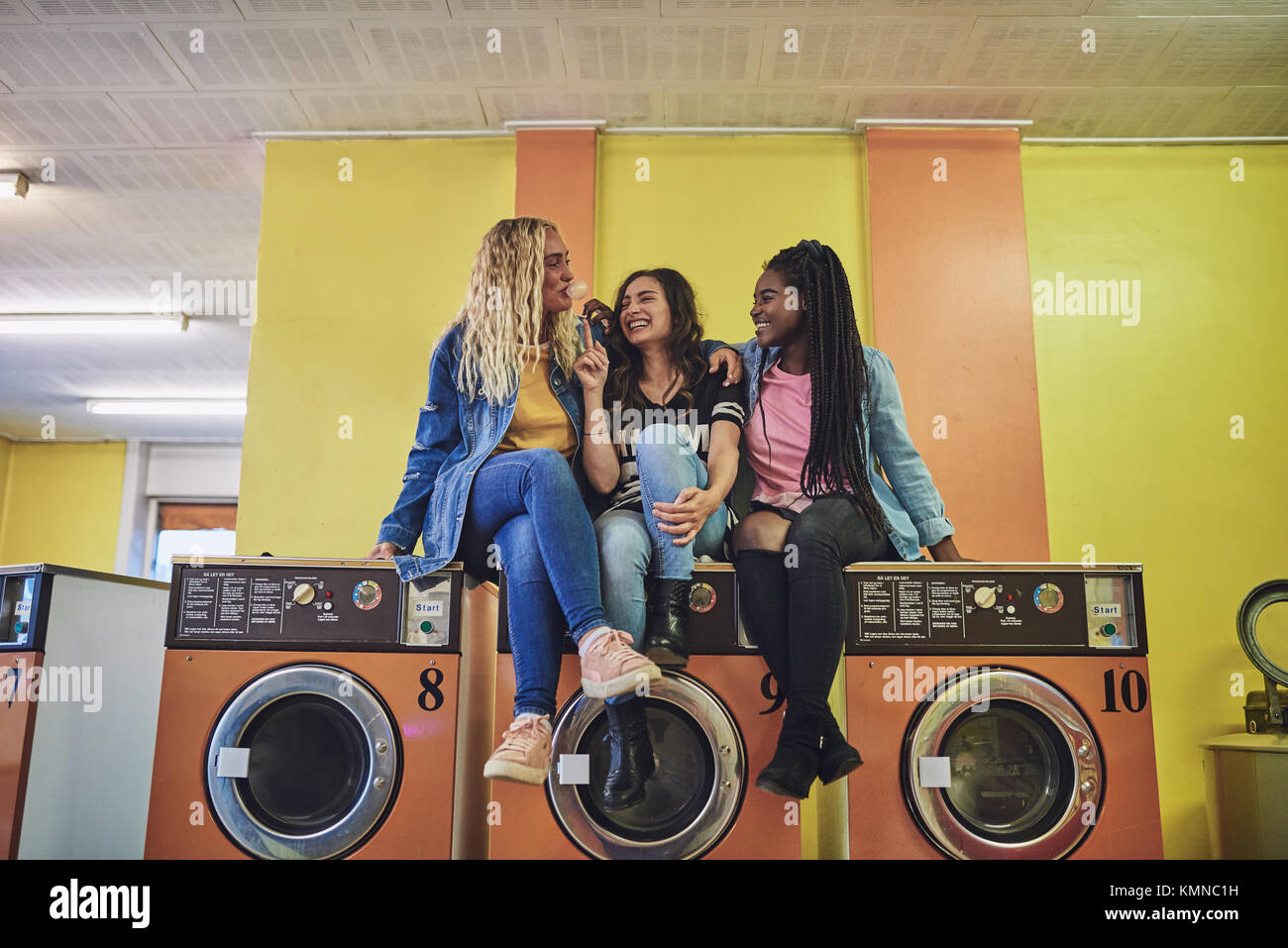Drei junge Freundinnen zusammen sitzen auf Waschmaschinen in einem  Waschsalon lachen und Seifenblasen mit Kaugummi Stockfotografie - Alamy