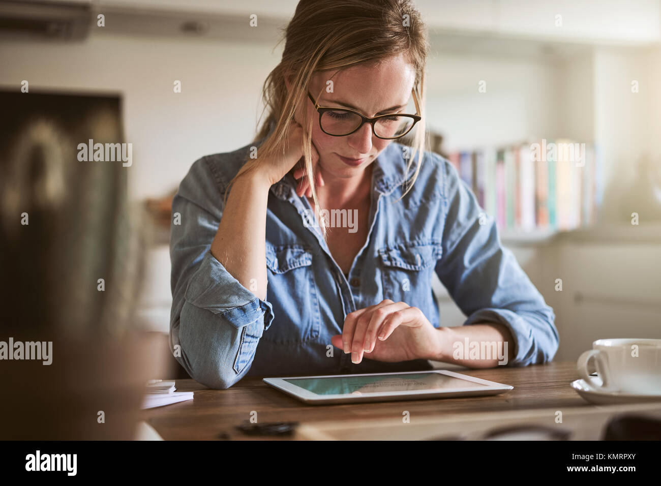 Junge Frau allein an ihrem Küchentisch, zu Hause zu sitzen, die online arbeiten mit einem digitalen Tablet konzentriert Stockfoto