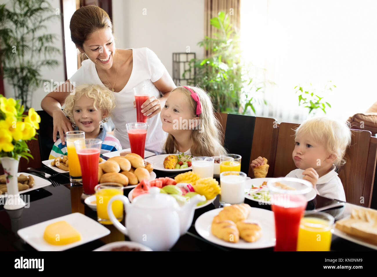 Gesunde Familie Frühstück zu Hause. Mutter und Kinder essen, tropische Obst, Toast, Brot, Käse und Wurst. Kinder trinken frisch gepressten Saft an sonnigen Stockfoto