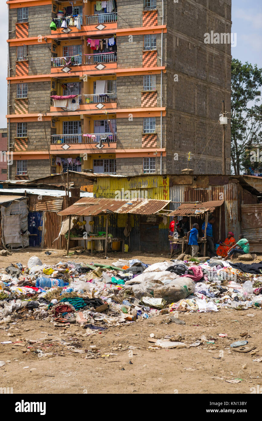Einen großen Haufen von Kunststoff- und allgemeinen Abfall Abfall liegt in der Mitte von einem offenen Bereich mit Menschen zu Fuß Vergangenheit, Wohnblocks und Hütten auf der Rückseite Stockfoto