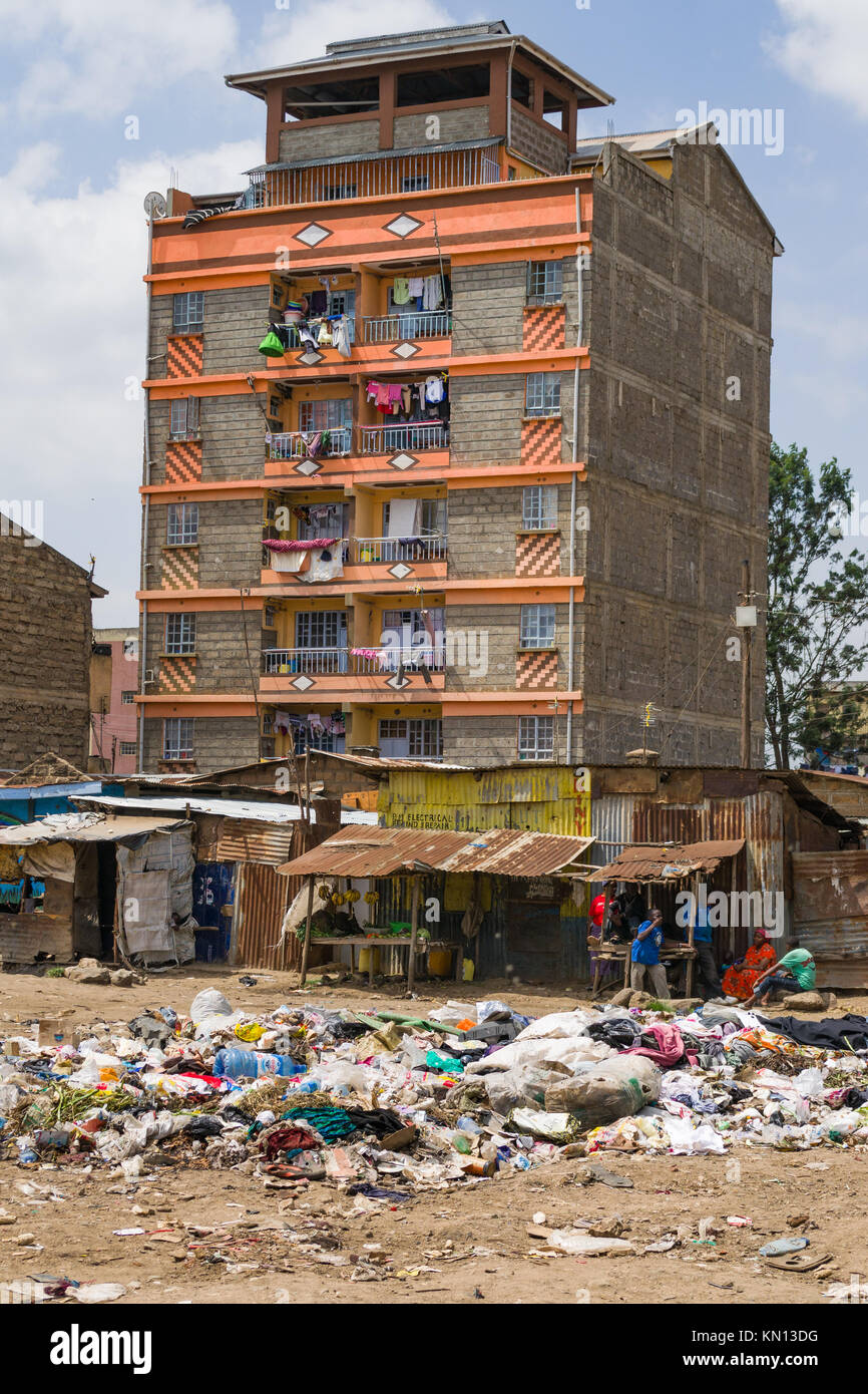 Einen großen Haufen von Kunststoff- und allgemeinen Abfall Abfall liegt in der Mitte von einem offenen Bereich mit Menschen zu Fuß Vergangenheit, Wohnblocks und Hütten auf der Rückseite Stockfoto
