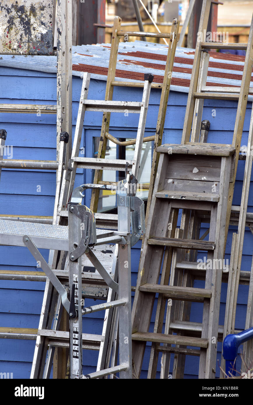Ein Stapel mit verschiedenen Leitern und Gruppen von Schritten aus, lehnte sich auf einem blauen Holzschuppen an einem alten Bootswerft neben einigen Gerüst in einem Bauherren Hof. Stockfoto