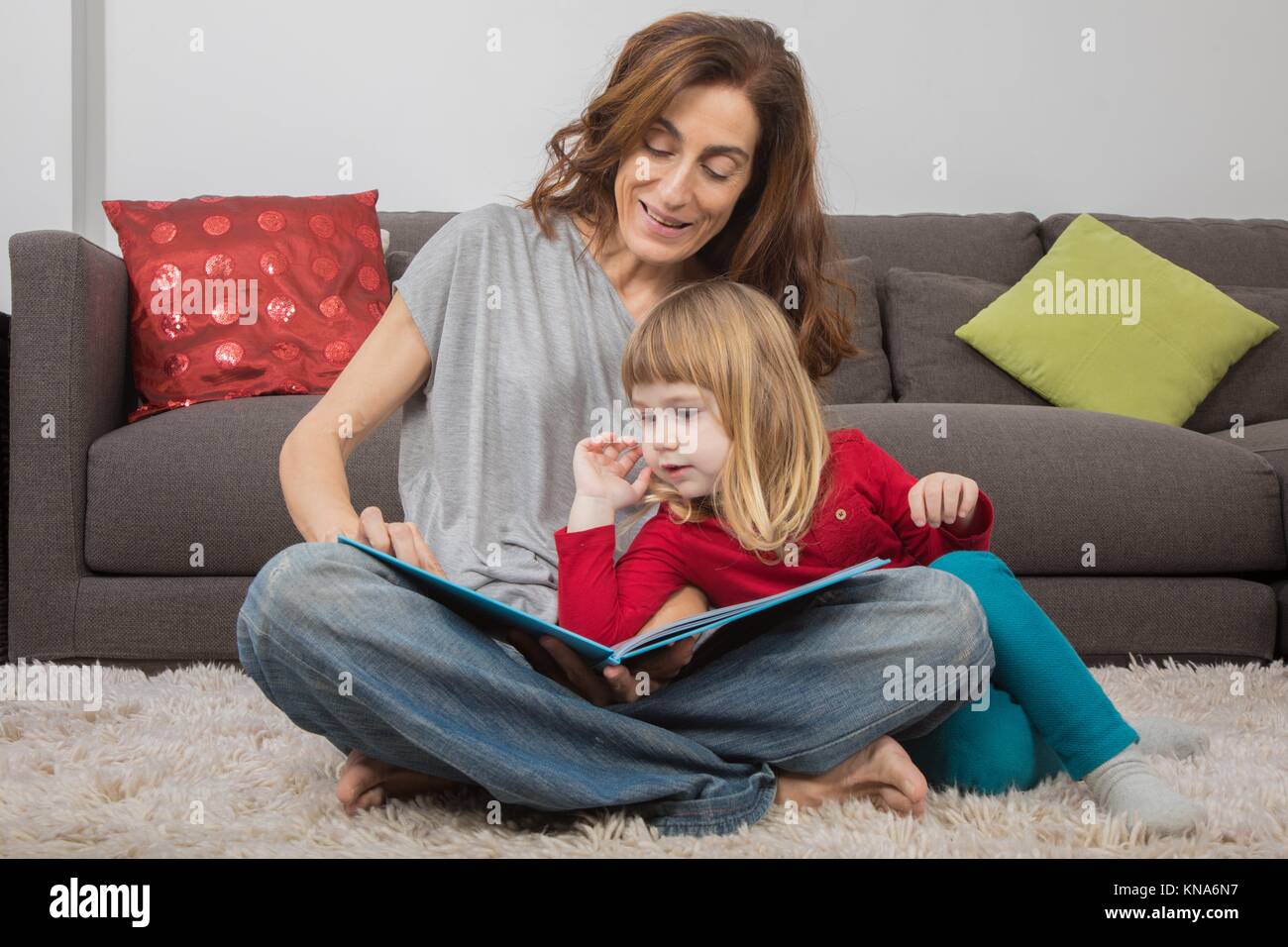 Blonde Kind drei Jahre alt, mit roten und grünen Kleidung, lehnte sich auf Mutter Frau in Jeans, zusammen eine Geschichte liest Märchen Buch, sitzen auf den Teppich Stockfoto