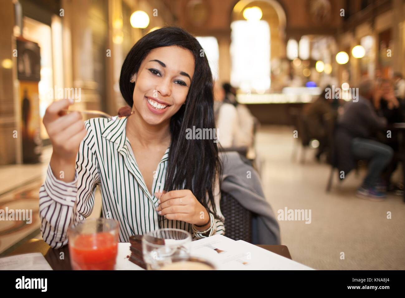 Junge Brünette Frau mit langen Haaren Kamera schauen und Lächeln während des Essens Schokoladenkuchen in Coffee-Shop. Stockfoto