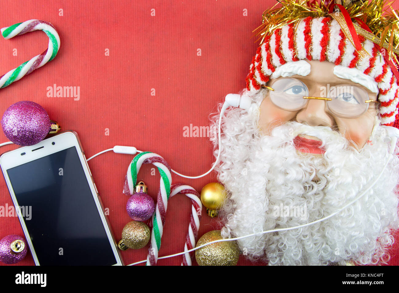 Santa mit Smartphone- und Ohrhörer, auf Rot mit Weihnachtsverzierungen Stockfoto