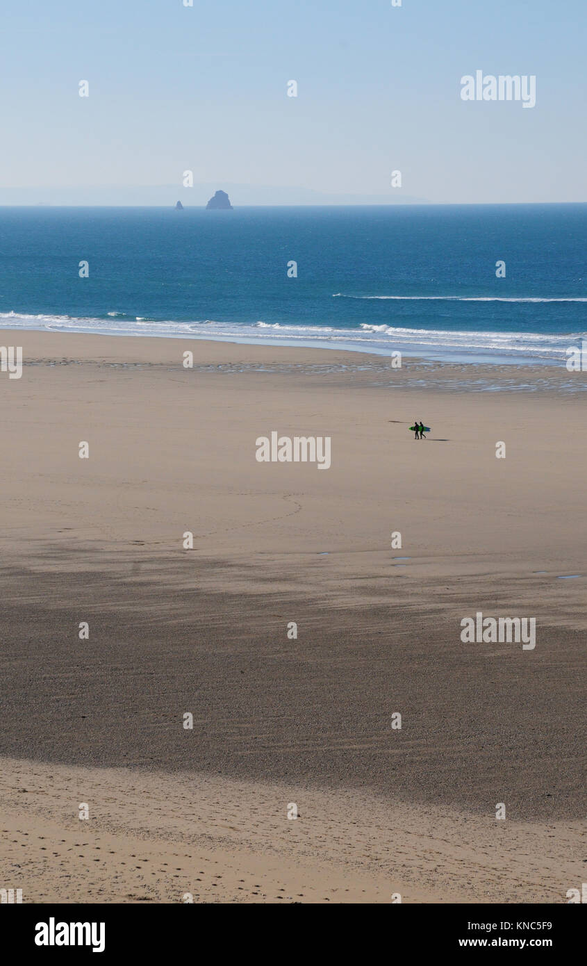 Zwei Surfer gehen über einen leeren Strand in Perranporth Cornwall. Bawden Felsen im Hintergrund Stockfoto Zwei Surfer gehen über einen leeren Strand in Perranporth Cornwall. Bawden Felsen im Hintergrund Stockfoto