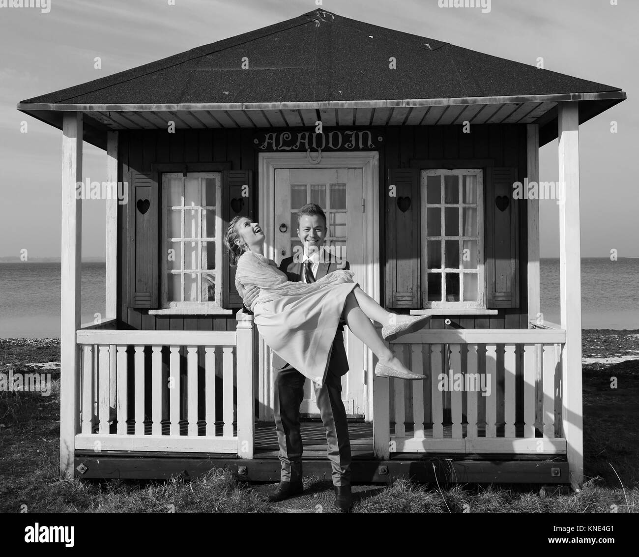 Attraktive glückliche junge Mann auf einem Knie vorgibt, schöne neue Frau auf der Werkbank zu durch gelbe Strandhütte für Spaß auf der dänischen Insel Aero vorschlagen Stockfoto