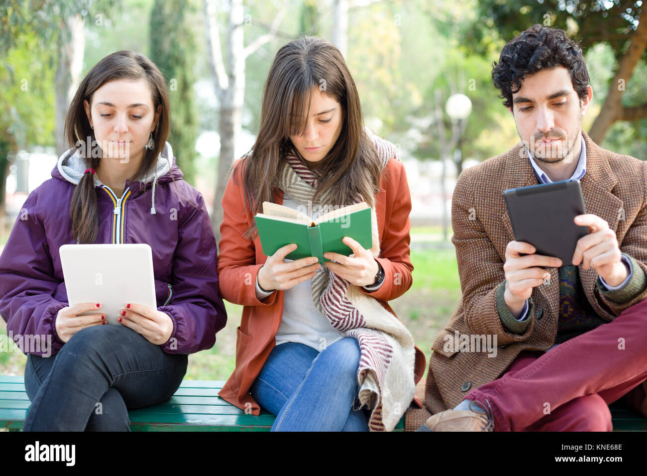 Eine Frau sitzen auf einer Bank in einem Park lesen Sie ein Buch aus Papier mit zwei Leuten Lesen ein E-Buch Stockfoto
