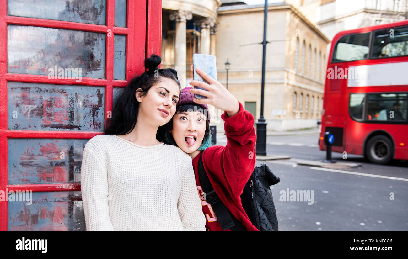 Zwei junge Frauen, die elegante Smartphone selfie durch rote Telefonzelle, London, UK Stockfoto