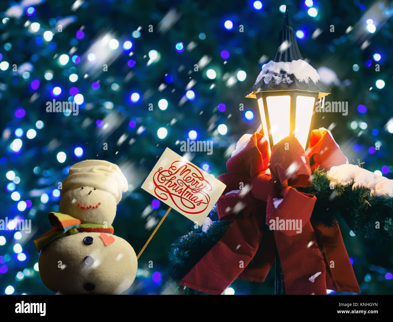 Schneemann Abbildung Holding ein Plakat. Text "Frohe Weihnachten!" darauf geschrieben. Straßenlaterne in Red Ribbon und Tannenzweigen gewickelt. Es schneit. Stockfoto
