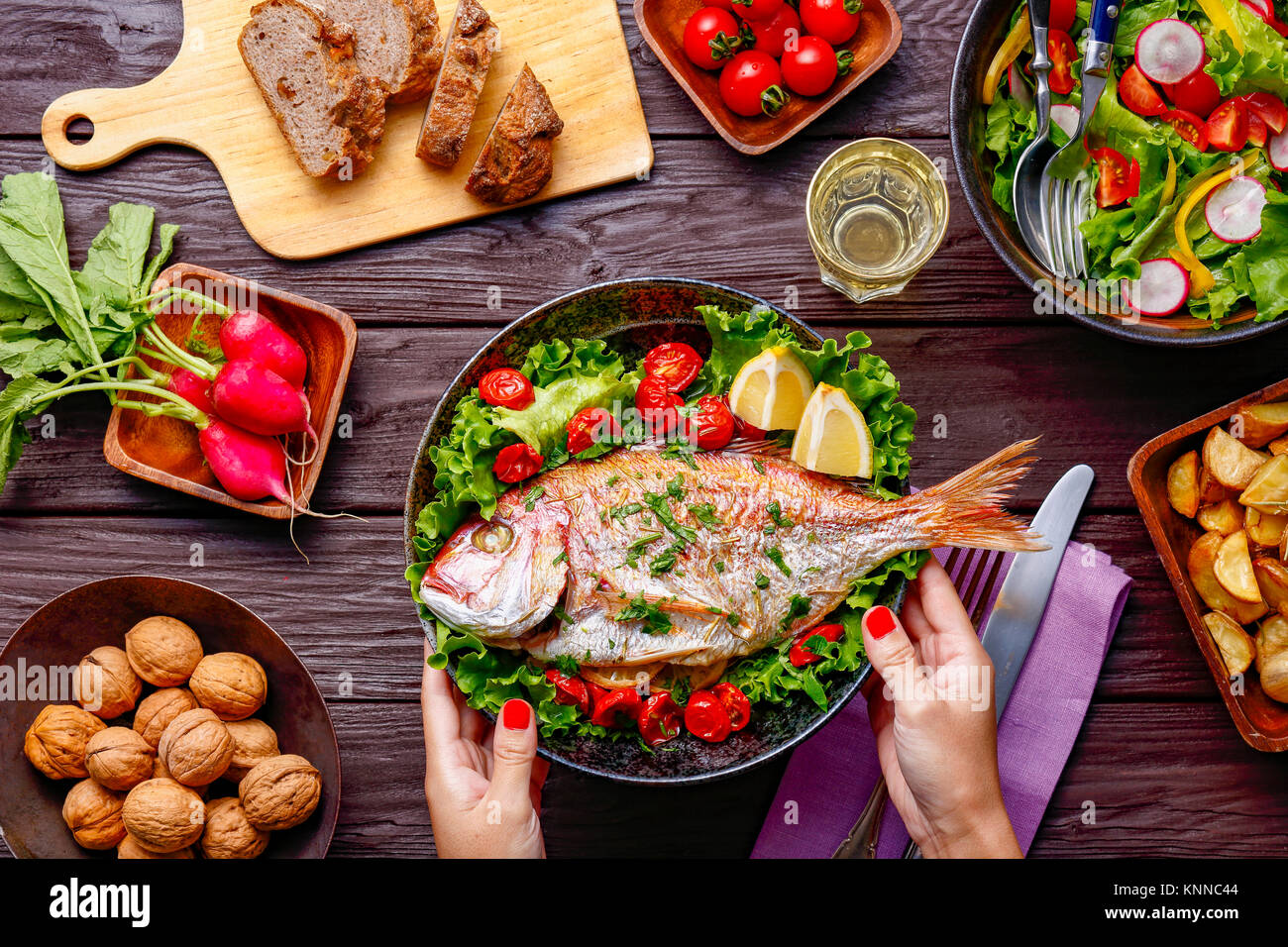 Frau Hand gegrillter Fisch am Tisch serviert mit farbenfrohen Speisen Stockfoto