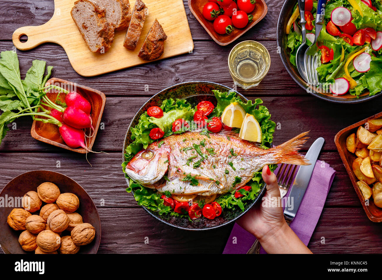 Frau Hand gegrillter Fisch am Tisch serviert mit farbenfrohen Speisen Stockfoto
