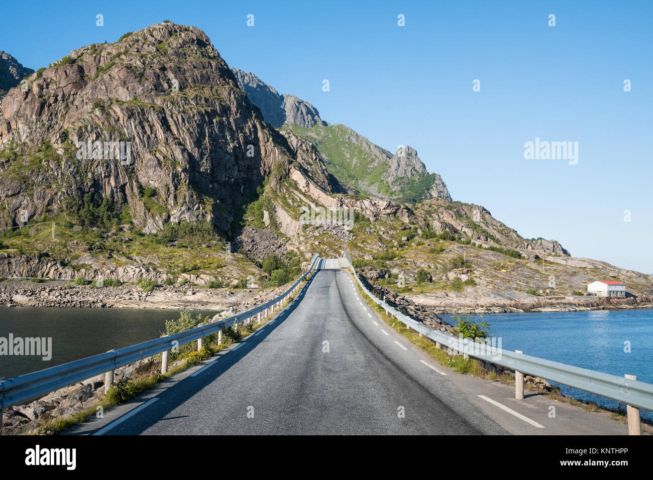 Gerade asphaltierte Straße mit Berg- und Meerblick gegen den blauen Himmel auf den Lofoten, Norwegen Stockfoto