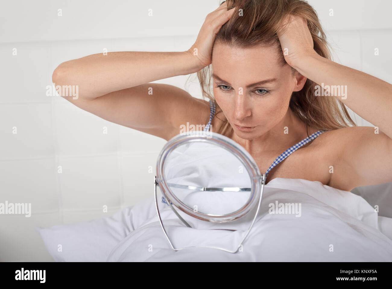 Ernsthafte junge Frau im Bett Blick auf sich selbst in einem kleinen tragbaren Spiegel auf dem Deckel zurück halten Ihre langen Haare mit den Händen ausgeglichen Stockfoto