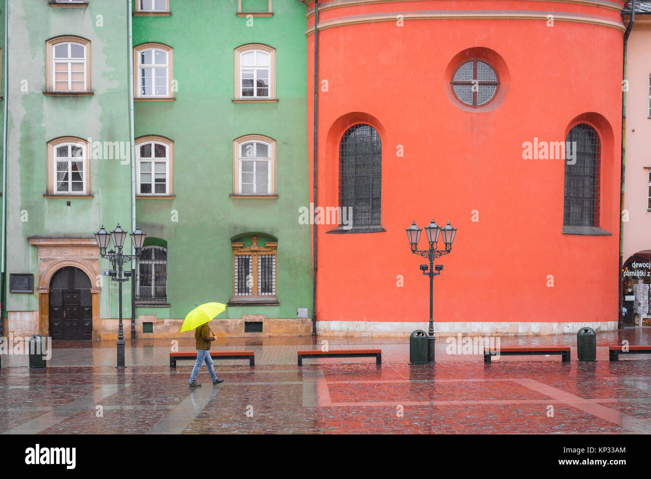 Man regen Stadt, ein Mann Unterschlupf unter einem gelben Regenschirm Wanderungen vorbei an einer Reihe von bunten Barockbauten in kleinen Marktplatz in Krakau, Polen. Stockfoto