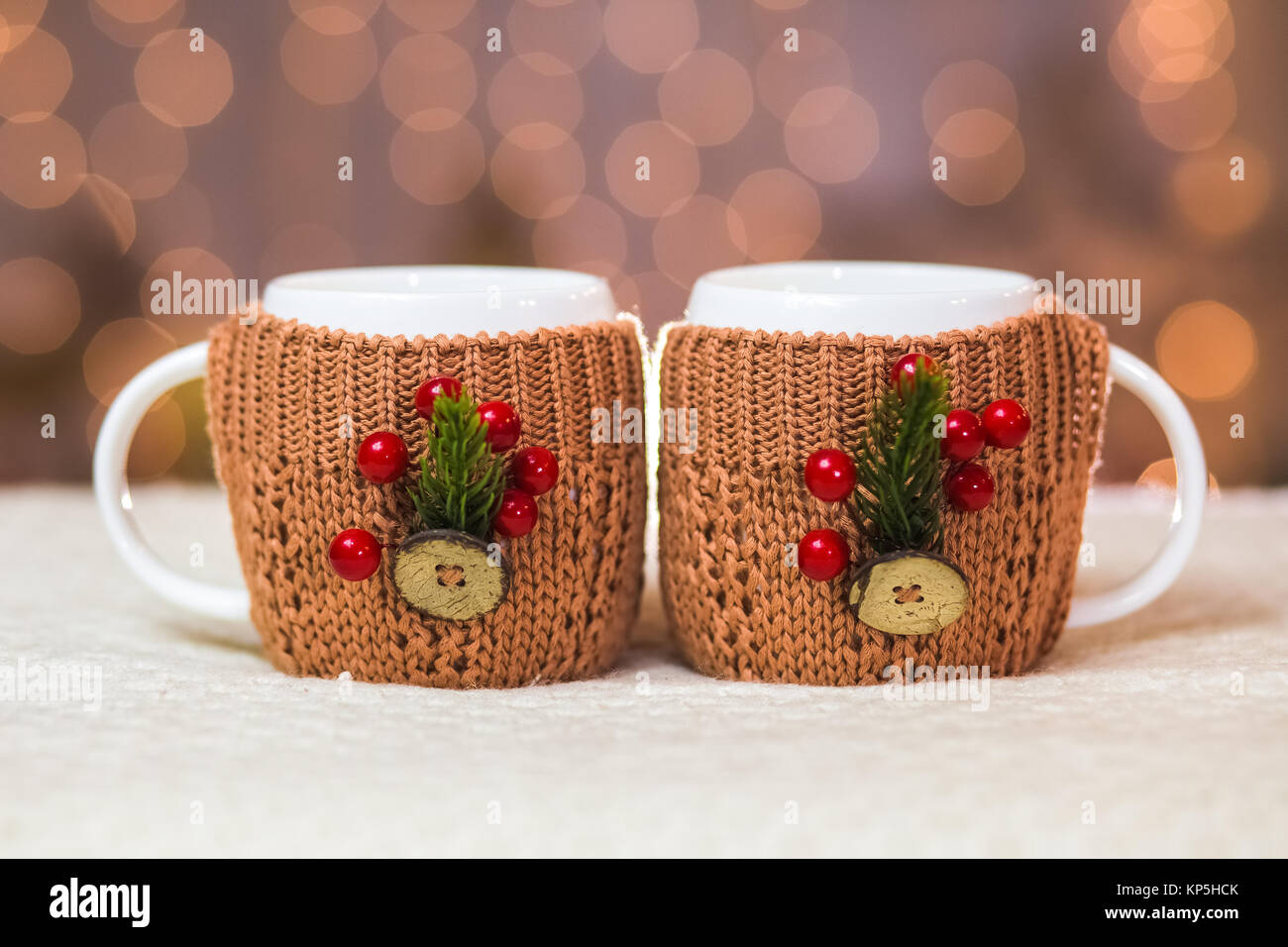 Nahaufnahme von zwei weißen Becher in süßen braunen gestrickte Pullover mit Weihnachtsschmuck an verschwommen Lichter von orange Girlande Hintergrund. Stockfoto