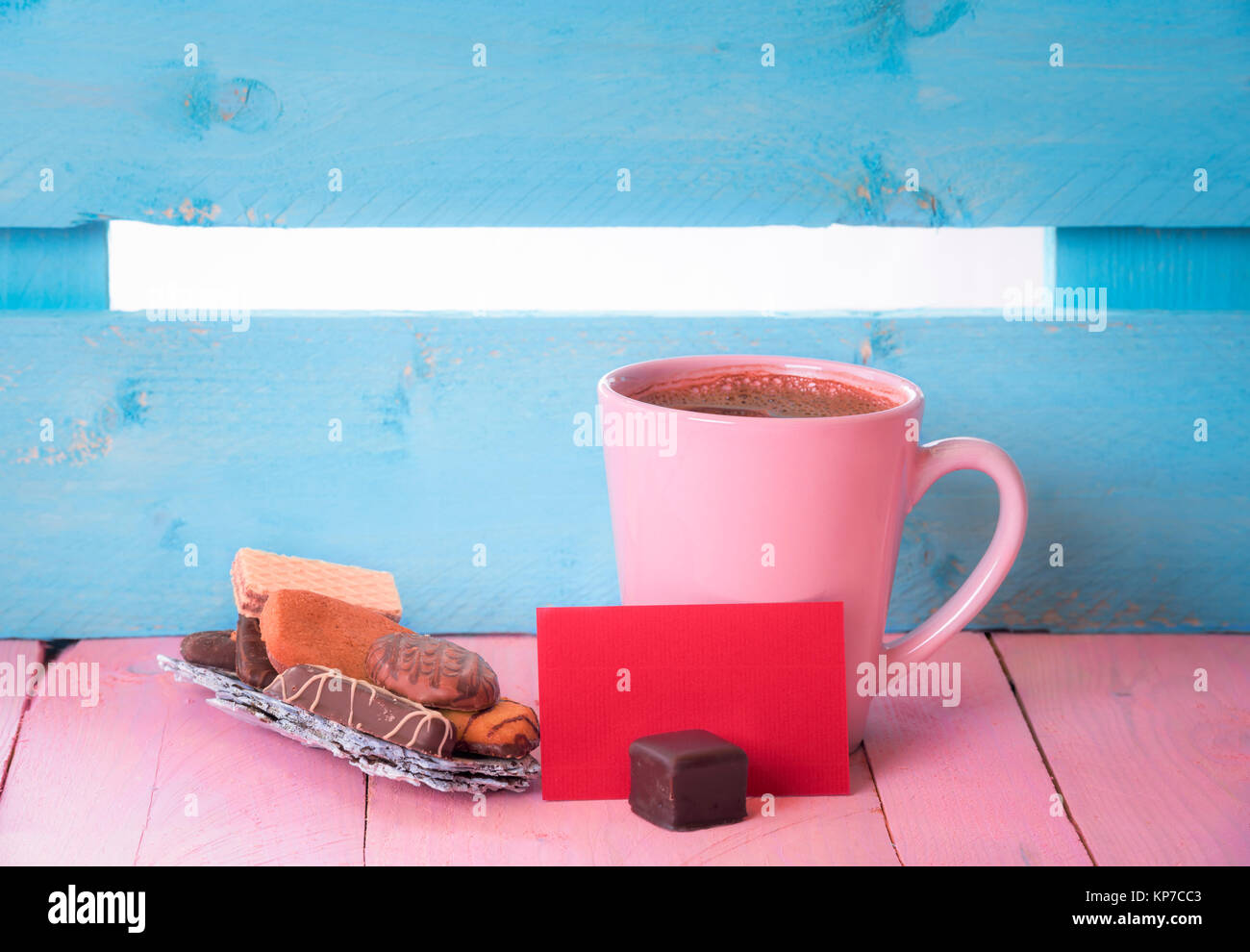 Tasse Kaffee in der Nähe von einem Haufen Süßigkeiten auf einem Baum Rinde und ein Leeres rotes Papier beachten Sie, auf einem rosa Holztisch und einem blauen Zaun im Hintergrund. Stockfoto