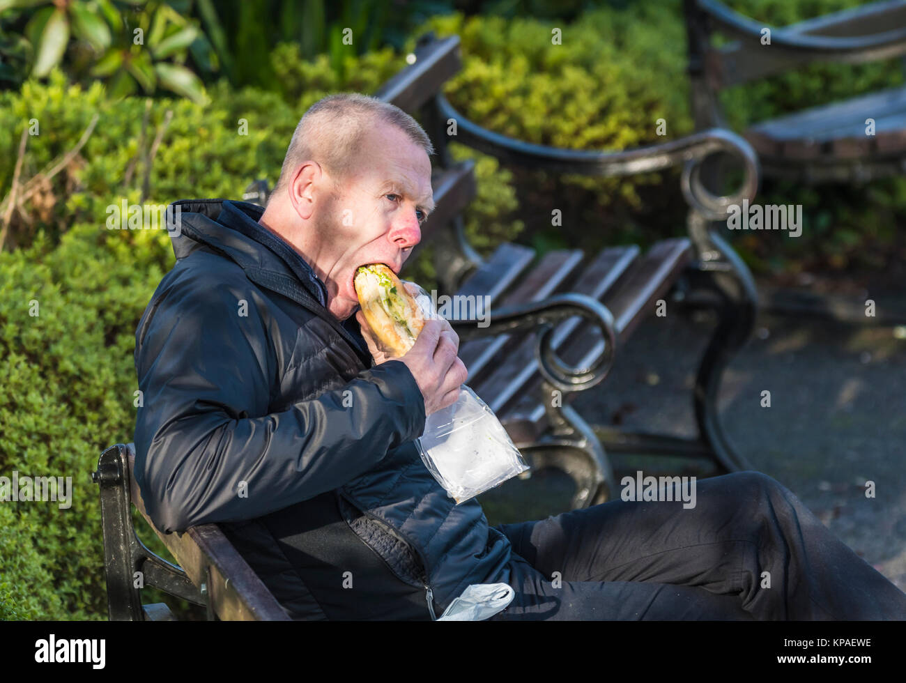 Man draußen sitzen Essen eine sub Sandwich (große lange Brötchen oder Baguette) für das Mittagessen, in Großbritannien. Stockfoto