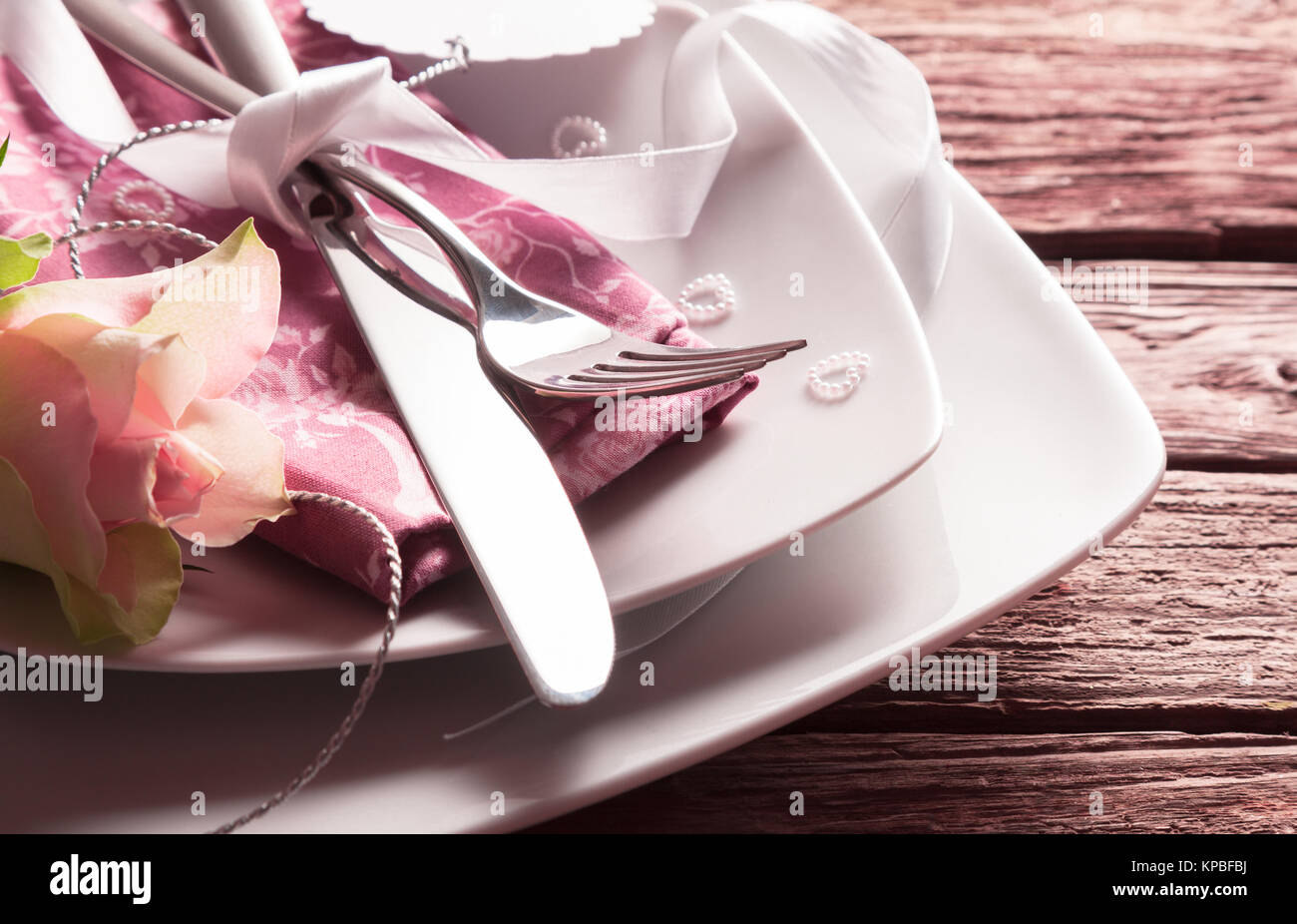 Nahaufnahme von romantische Hochzeit oder zum Muttertag Ort Einstellung - Messer und Gabel gebunden mit weißem Band auf Rosa Serviette auf Weiße Platten mit rosa Rose und winzige Perle Herz auf rustikalen Holztisch Stockfoto